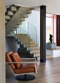 Leather armchair with modern, metal frame in open-plan stairwell with entrance area and black metal pillar in front of stone staircase with glass balustrade