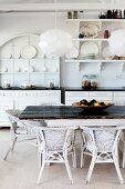 Modern pendant lamps above rustic table and white rattan chairs in front of kitchen counter and white crockery on wall-mounted shelves