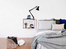Innen weiss gestrichene Holzkiste mit Klemmleuchte und Deko als Nachttisch an die Wand gehängt, daneben ungemachtes Bett und Wäschekorb