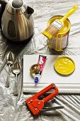 Pot of yellow paint, stapler, white skirting board and dessert spoons