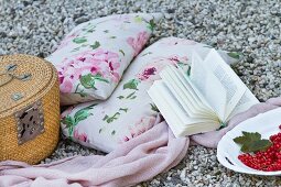 Cushions with a floral pattern, a blanket, a book and a picnic basket on a gravelly surface