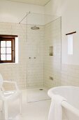 Partially visible white Ghost chair and bathtub in front of floor-level shower with glass partition in bathroom with white wall tiles