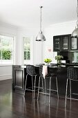 Elegant, black fitted kitchen with counter and bar stools on fine, dark parquet floor; white bay window and chrome pendant lamps