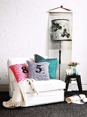 White upholstered chair with various scatter cushions next to vase of flowers on black stool, wall hanging with standard lamp motif on white wall