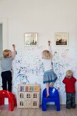 Drei Kinder mit Plastikstühlen und Spielhaus beim Bekritzeln einer weissen Wand mit gerahmten Fotografien