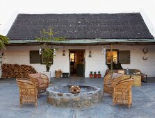 Wicker armchairs on stone floor around circular stone fire pit with simple country house in background