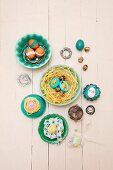 Various Easter eggs on nests of straw, bent wire and ribbon in bowls on pale wooden surface