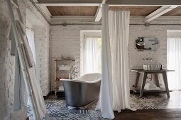 Free-standing vintage bathtub and washstand screened by curtains, whitewashed brick walls and grey and white patterned tiled floor
