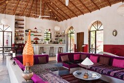 Lounge area with colourful seat cushions on masonry benches in open-plan interior with palm thatch roof