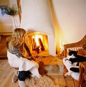 Frau vor offenem Kaminfeuer und Katze auf Korbstuhl