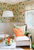 Wicker chair, standard lamp and side table against pale, wood-panelled dado and floral wallpaper above dado rail; orange and green accessories and flowers