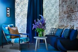 Blue painted wall, patterned wallpaper and brick wall as backdrop for blue seating area with sofa, armchair and bouquet of lupins on side table