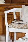 Gebeizter Holzschrank hinter weiss lackiertem Armlehnstuhl; Notizbuch mit Brille auf gemustertem Sitzkissen