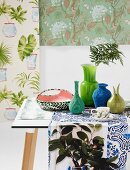 Collection of vases in shades of green and blue arranged on runner in front of various samples of floral wallpaper