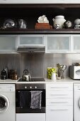 White fitted kitchen with wall units and various glass vessels and collectors' items on dark wooden shelf