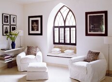 Armchairs and footstool with white loose covers in corner of room in front of Gothic window niche with window seat