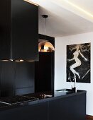 Black kitchen counter with sink and hob below extractor hood next to pendant lamp with copper-coloured lampshade