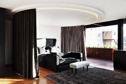 Bed and couch below circular ceiling element with integrated curtains and lights next to floor-to-ceiling window