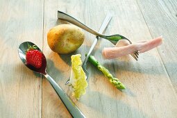 A still life featuring a strawberry, a potato, a lettuce leaf, an asparagus stalk and ham on spoons and forks
