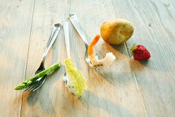 A still life featuring an asparagus stalk, a salad leaf, a prawn, a potato and a strawberry on forks