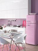 White kitchen with lilac wall and pink retro fridge; purple and orange rug below glass table