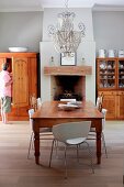 Pale grey room with open fireplace and antique dining table below elaborate chandelier; white designer chairs and antique wooden chairs around table