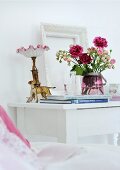 Rosafarbener Rosenstrauß mit antiker Nachttischlampe und Hundefigur auf weiß lackiertem Nachttisch