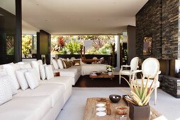 White sofa combination with scatter cushions and rustic low coffee table in front of Neo-Rococo chairs in spacious living room with open fireplace in grey stone wall