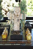 Necklaces hung on cross-shaped jewellery stand behind black box, bottles of perfume and necklaces laid out on surface