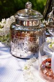 A silver jar, white lilac flowers and cakes on a table in the garden