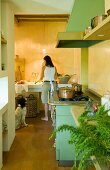 Copper pans on matt green, vintage gas cooker in country-house kitchen; dog sitting next to woman cleaning vegetables in long sink