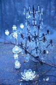 Glass candlesticks of various sizes and metal candelabra with crystal pendants on wooden deck next to lake