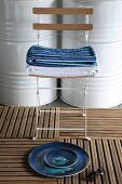 Still-life arrangement of plates on decking tiles in front of stacked towels on folding chair and white-painted metal barrels