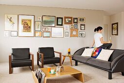 Modern coffee table in pale wood and armchairs with black leather covers in front of gallery of pictures on wall; woman sitting on back of grey sofa