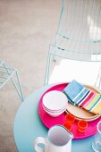 Pink tray of crockery on round blue terrace table and delicate wire mesh chairs in background