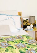 Teilweise sichtbares Bett mit geblümter Tagesdecke und Kissenstapel neben Nachttisch mit Kaffeetasse vor Tischleuchte