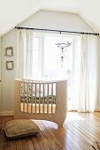 Custom-made, pale wood cot in front of balcony doors with airy curtains in attic nursery