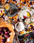 Verschiedene Schalen aus verschiedenen Materialien, teilweise bemalt mit Vogelmotiven und getrocknete Blumen auf Stoff mit Herbstlaub Motiv
