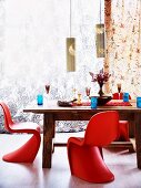 Rote Panton Stühle um massiven Holztisch mit blauen Trinkgläsern gedeckt und blumige zarte Vorhänge im Hintergrund