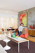 White stool, delicate coffee table and woman sitting on salmon-pink armchair on rug in front of modern artwork on exposed concrete wall; dining area next to glass wall