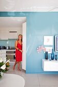 Interior painted pale blue with partially visible floating sideboard with collection of blue vases; woman wearing red dress in open doorway