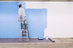 Mann steht auf Leiter und streicht Wand mit blauer Farbe