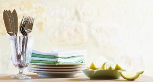 Cutlery in a glass, stacked plates and napkins; lemon wedges to one side