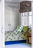 Bed with patterned bedspread and stacked scatter pillows in front of fitted wardrobe in white, wood-panelled bedroom