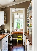 Country-house-style galley kitchen with view of writing desk and yellow stool through doorway