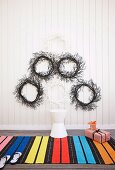 Wreaths in black and white on a white wooden wall, in front of it a colorful striped carpet with a stool and Christmas presents