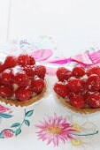 Two strawberry tartlets on a floral tablecloth with plastic cutlery in the background