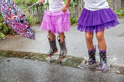 Two girls in purple tulle skirts and colourful wellington boots jumping in puddle