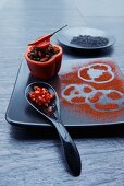 A stuffed pepper filled with wild rice, chilli and tomatoes with a spoonful of chilli rings and a black plate with paper prints in paprika powder