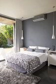Elegant double bed with grey upholstery against dark grey wall and between two white, designer pendant lamps; view of planted terrace